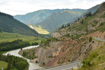 Naklejka premium A fragment of an asphalt road running along the slope of a high mountain and a winding river in a valley on a cloudy summer day.
