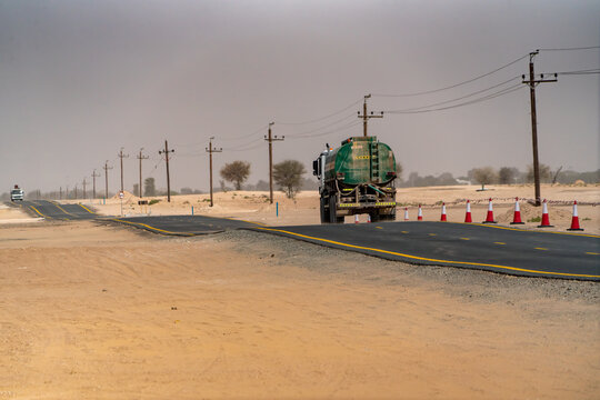 View of a green water tanker truck driving on a newly paved asphalt road through the desert landscape with utility poles and traffic cones Dubai, Dubai, United Arab Emirates.