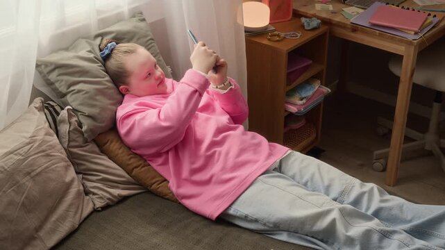 High angle handheld shot of teenage Caucasian girl with Down syndrome lying on bed and looking at smartphone screen, scrolling news or watching videos in cozy room