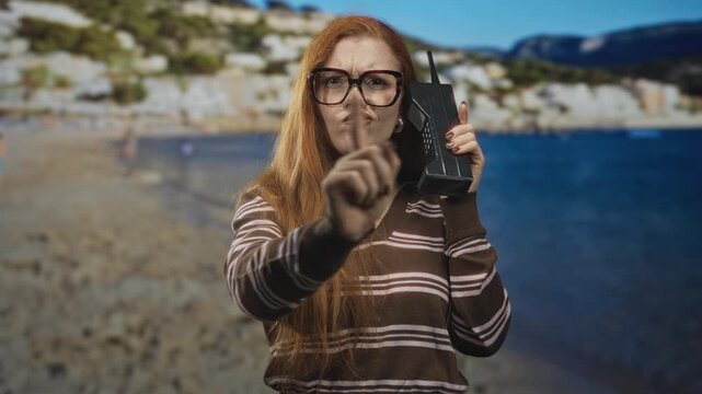 Redhead woman holding retro brick cellphone and pointing finger toward camera in studio beach set; defiance.