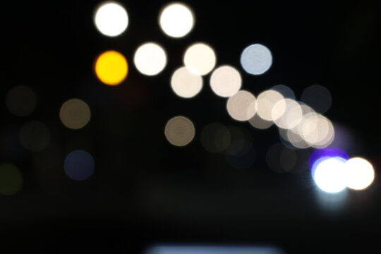 Night street glows with blurred lights and motion. Cars pass, leaving trails of brightness in the dark. Trees and signs softly fade into bokeh behind. Urban life hums under a silent, starless sky