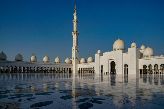 View of the Sheikh Zayed Grand Mosque featuring white marble domes, a tall minaret, and a reflective floral courtyard under a clear blue sky in Abu Dhabi,United Arab Emirates.