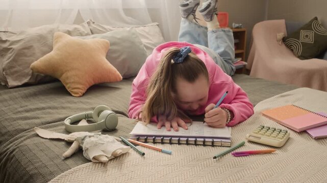 Handheld shot of focused teenage Caucasian girl with Down syndrome lying on bed surrounded by educational materials and doing homework