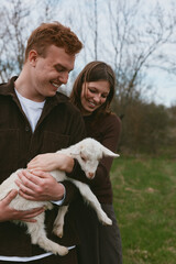 Naklejka premium A joyful couple outdoors holds a fluffy lamb, smiling as they cradle the young sheep in a green field, capturing a candid lifestyle moment of gentle animal care and love