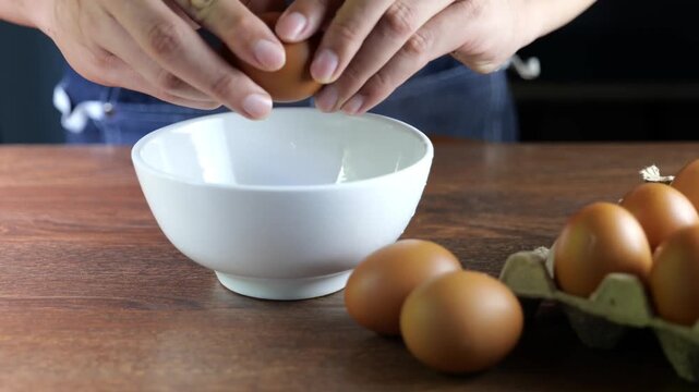 Close-up footage of hands cracking a fresh brown egg into a white ceramic bowl on a wooden table, with eggs in a carton nearby. Perfect for cooking, baking, breakfast, and food preparation concepts.