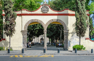 Fototapeta premium Arco de entrada colonial al atrio de San Juan Bautista en Coyoacán, Ciudad de México, con ambiente de parque