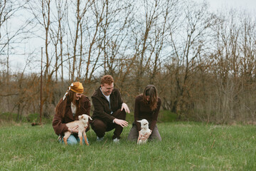 Naklejka premium Three people in casual autumn clothing interact with two goats in a grassy park, capturing a warm lifestyle moment outdoors, highlighting family, friends, nature, and animals in a tranquil field
