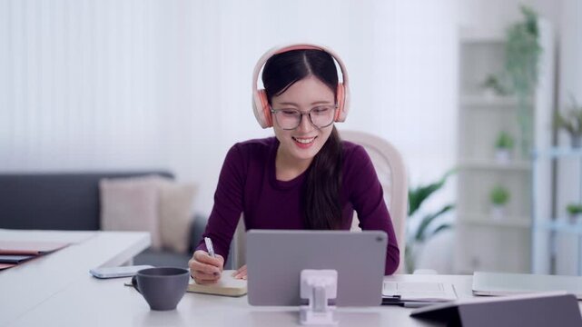 young Asian woman student wearing headphones glasses using tablet taking notes studying online learning video conference remote work from home office desk with
