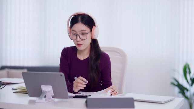 young Asian businesswoman holding credit card using tablet computer for online shopping payment working remote from modern home office wearing pink headphones