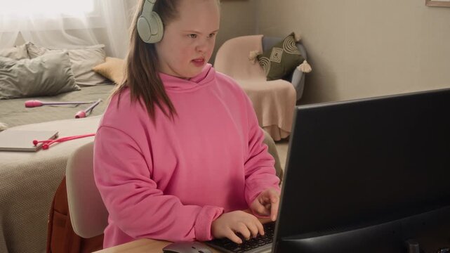 Medium shot of cheerful teenage Caucasian girl with Down syndrome wearing headphones sitting at desk in front of computer and having fun while playing videogame in cozy room