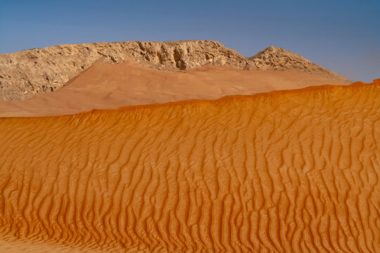 View of red sand dunes with wind ripples and a rocky mountain ridge under a clear blue sky in the desert Dubai, Dubai, United Arab Emirates.