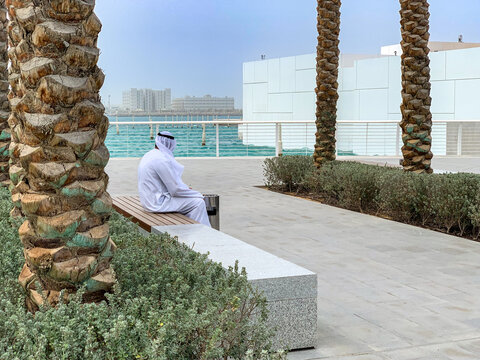 View of a man in traditional Emirati clothing sitting on a bench along a waterfront promenade with palm trees and modern buildings in Dubai, Dubai, United Arab Emirates.