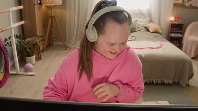 Chest up shot of teenage Caucasian girl with Down syndrome in headphones sitting at desk in cozy bedroom and enjoying playing videogame or chatting with friends using computer
