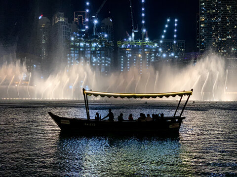 View of the Dubai Fountain at night with a traditional abra boat carrying people against a backdrop of illuminated skyscrapers and construction cranes in Dubai, Dubai, United Arab Emirates.