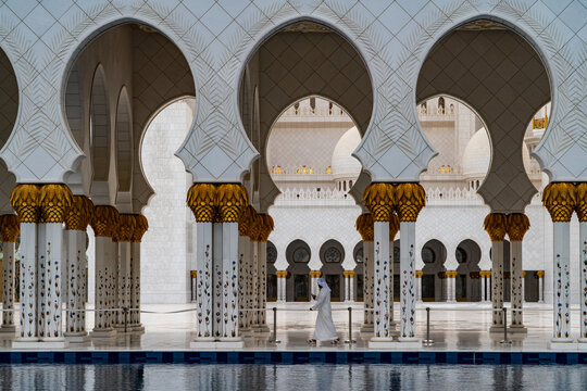 View of the white marble arches and intricate floral patterns of a grand mosque with a man walking through the courtyard in Abu Dhabi,, Abu Dhabi,, United Arab Emirates.