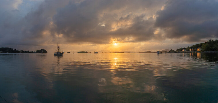 View of fishing boats anchored in a calm bay under a dramatic sunset with heavy clouds and golden light reflecting on the water United States.