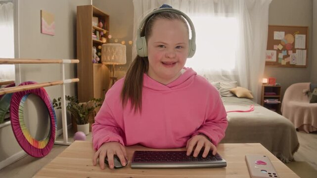 Medium POV shot of focused teenage Caucasian girl with Down syndrome wearing headphones sitting at desk in cozy bedroom in front of computer and playing videogame using keyboard