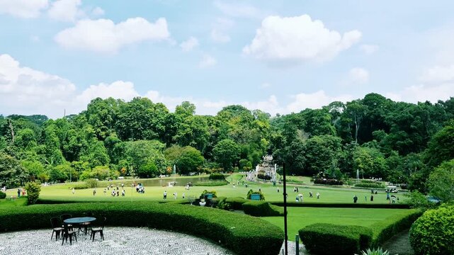 Wide view of Bogor Botanical Gardens with visitors on the central lawn, a distant fountain, and dense tropical trees under bright daylight.