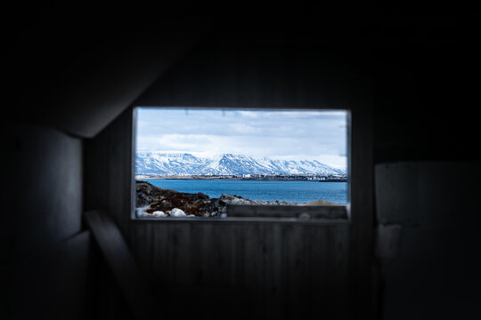 View of the interior of an abandoned farmhouse looking out through a window at snow-capped mountains and the blue sea in Hafnarfjordur, Hafnarfjardarkaupstad, Iceland.