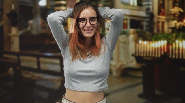 Woman redhead wearing glasses and gray crop top showing bare midriff with hands behind head in church interior near votive candles; playful confidence.
