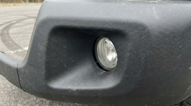 Close-up of a circular fog light assembly integrated into a black textured plastic front bumper trim of a vehicle, highlighting the pebbled surface detail and clear glass lens.