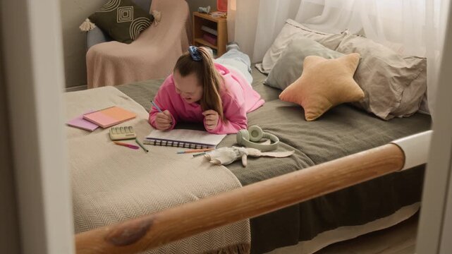 High angle full shot of teenage Caucasian girl with Down syndrome studying on bed in cozy room surrounded by educational materials and calculator