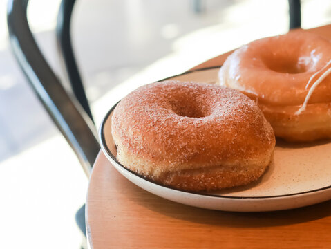 Two classic yeast doughnuts on a light-colored, speckled plate, one generously coated in cinnamon sugar and the other with a simple sugar glaze, resting on a wooden cafe table near a window.