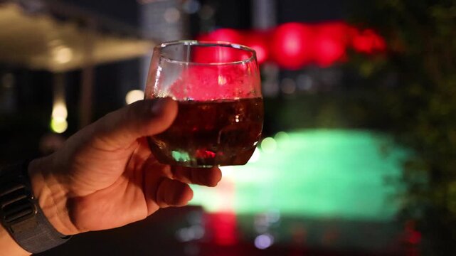 Two people clinking glasses with dark cocktail drinks at night rooftop party by swimming pool.