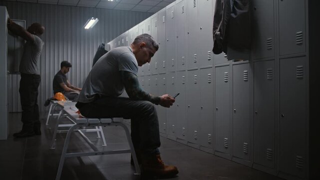 Tattooed Male Worker Sits on Grey Bench in Modern Industrial Locker Room, Using Smartphone to Make Call While Coworkers Undress After Factory Shift. Concept of Connecting With Home During Break.