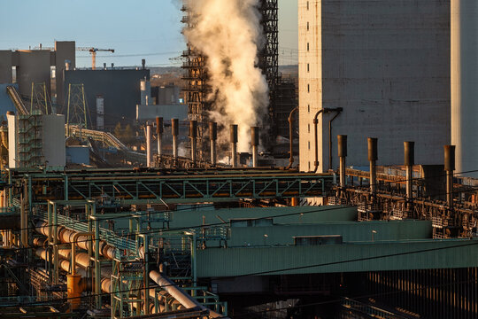 View of a massive industrial steel plant with thick white smoke rising from chimneys and a complex network of pipes in Duisburg, North Rhine-Westphalia, Germany.