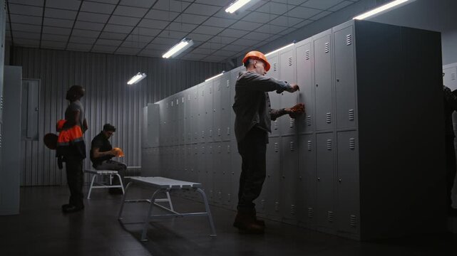 Group of Blue Collar Professionals in Orange Safety Helmets Talking and Opening Metal Lockers in Clean Industrial Dressing Room After Finishing Duty. Concept of Teamwork and Work Life Balance.