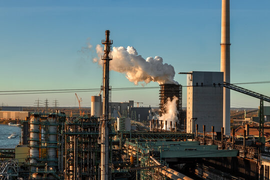 View of an industrial steel plant with smoking chimneys and complex piping systems under a clear sky during sunset Duisburg, North Rhine-Westphalia, Germany.