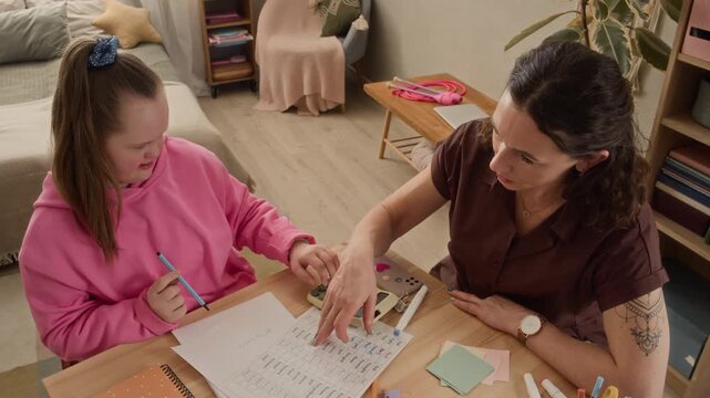 High angle shot of teenage Caucasian girl with Down syndrome doing math homework with her mothers supervision, using calculator while studying at desk in cozy bedroom