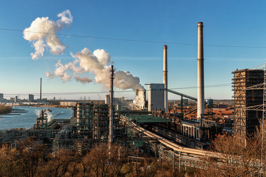 View of an industrial plant with smoking chimneys and steel structures along the Rhine river under a clear blue sky at sunset in Duisburg, North Rhine-Westphalia, Germany.