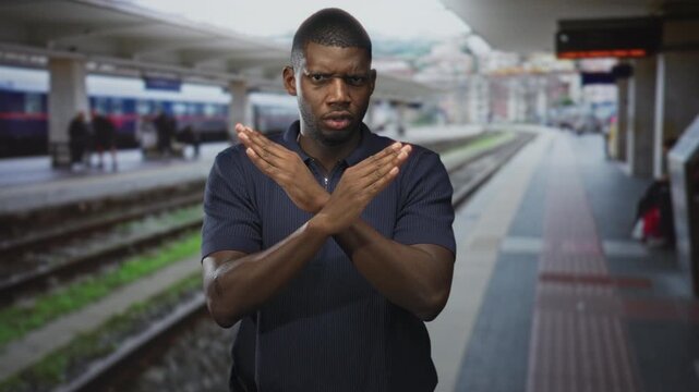 Man with crossed arms hands forming x gesture on a station platform inside a building beside train tracks and tactile paving; defiance.