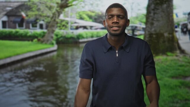 Man pinches nose with hand on canal side street, wearing glasses and dark polo near a tree lined canal; disgust.