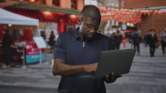 Young black man wearing glasses typing on laptop, hands visible in busy street market; concentration productivity focus.