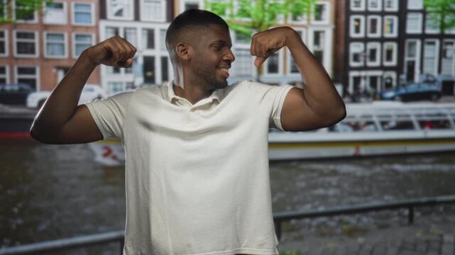 Man flexes biceps and points to arm on a street by a canal with boat and row of buildings; strength success confidence.