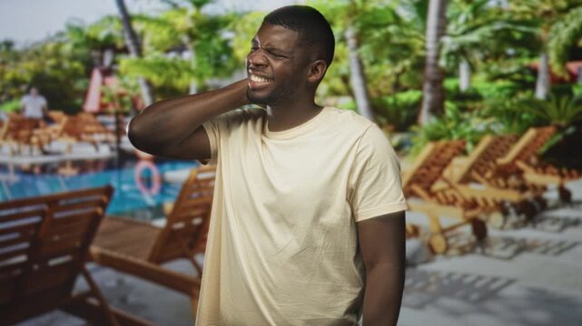 Man hand on neck wincing with neckache against a studio pool backdrop with loungechairs and palms; vacation discomfort.