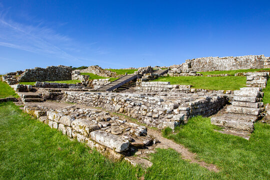 View of the ancient Roman fort ruins of Vindolanda with stone walls, green grassy slopes, and wooden stairs under a clear blue sky Hexham, England, United Kingdom.