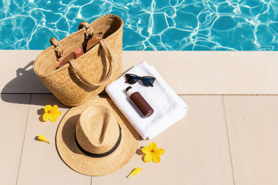 Straw bag with sun protection items rests on pool deck. Includes hat sunglasses sunblock lotion white towel yellow flowers. Relaxing summer day at blue water.