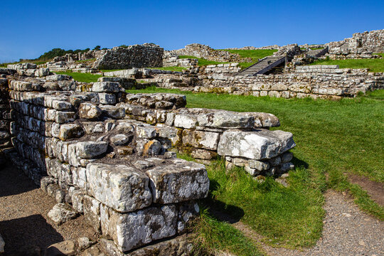 View of the ancient Roman ruins of Vindolanda featuring stone walls and grassy foundations under a clear blue sky in Hexham, England, United Kingdom.