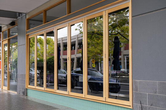 Modern retail shopfront with large glass folding doors and timber frames. Stylish cafe or restaurant exterior with wooden bi-fold glass windows along a paved public walkway