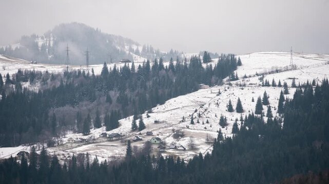 Time lapse of snowy mountain village with drifting fog over forested hills. Winter landscape reveals rural houses, pine trees, and changing weather in highland countryside