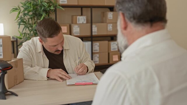 Man pointing at clipboard and signing a parcel in a building with stacked boxes and handshake gesture; trust agreement.