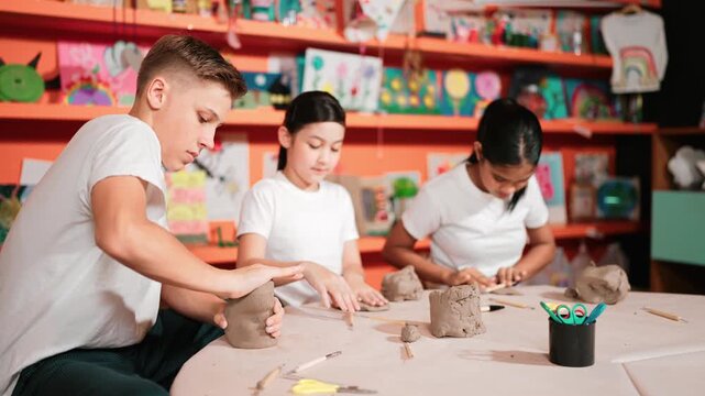 Caucasian smart teenager modeling clay while craving glass of clay at art lesson. Group of multicultural happy student working at dough in pottery workshop. Creative activity concept. Edification