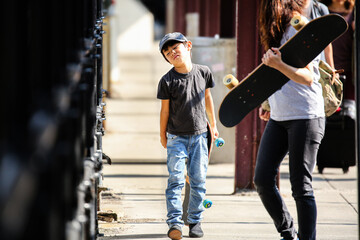 A young boy looking tired and slightly grumpy while holding a skateboard on a street in Boston during a long family trip. © Kenji