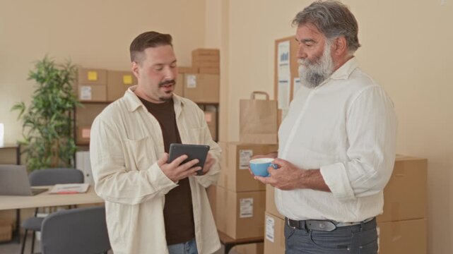 Man holding a tablet while colleague holds a coffee cup and they discuss stacked packages and shipping labels in an office building; teamwork efficiency.
