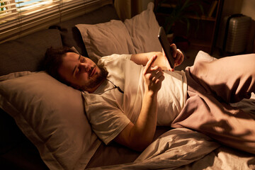 Fototapeta premium Young adult Caucasian man lying in bed, using smartphone at night, scrolling before sleep and showing screen time habit. Useful for sleep disruption, mobile addiction, bedtime routine themes
