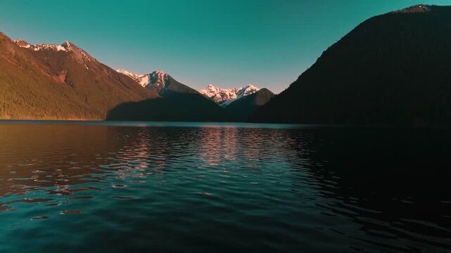 Peaceful dark lake surface rippling gently while reflecting deep teal twilight sky stretching toward distant rugged mountain peaks brightly illuminated from golden sunset approaching aerial drone